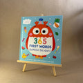 Children's book titled '365 First Words' with an owl illustration on a stand against a brown background