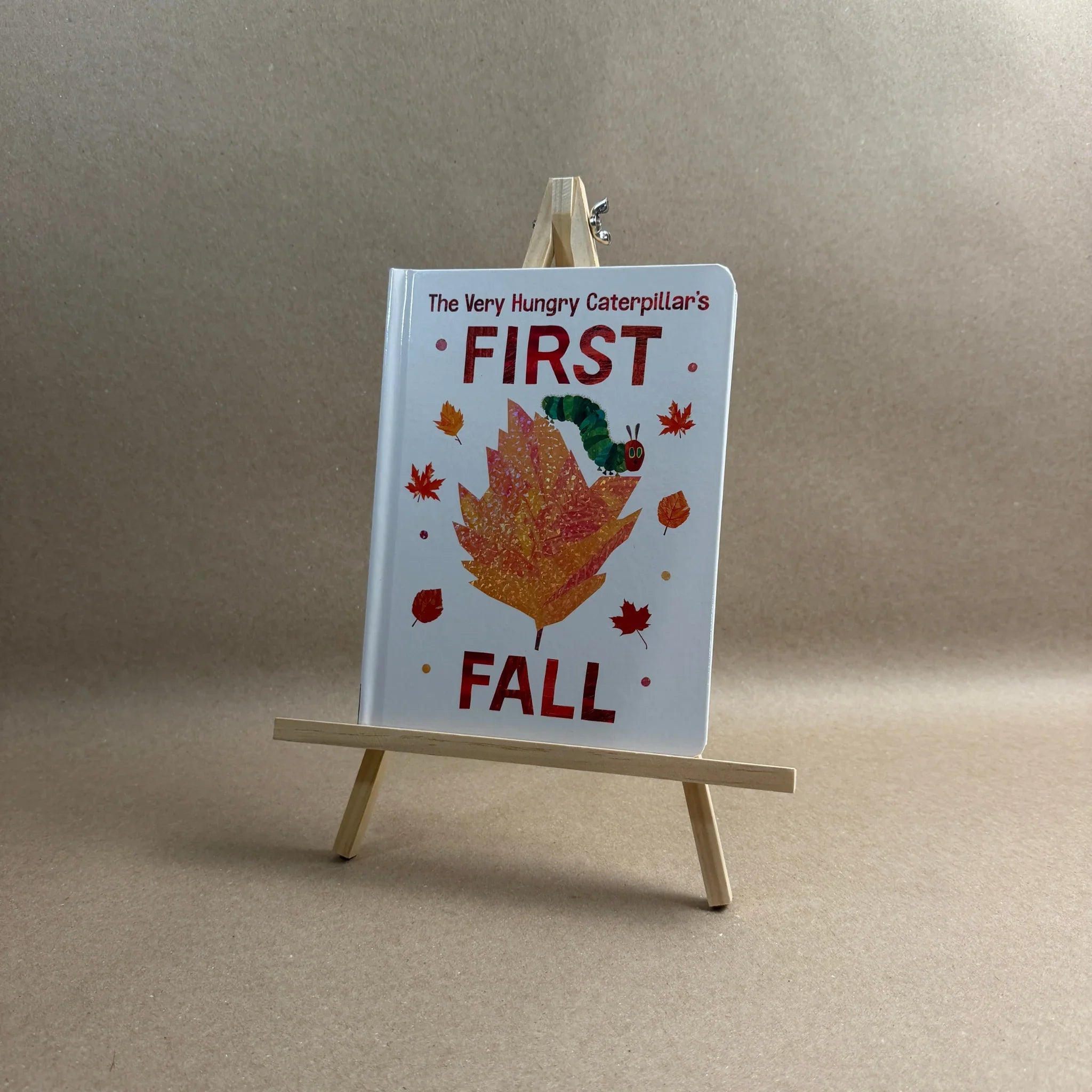 Children's book titled 'The Very Hungry Caterpillar's First Fall' on a small wooden easel against a neutral background