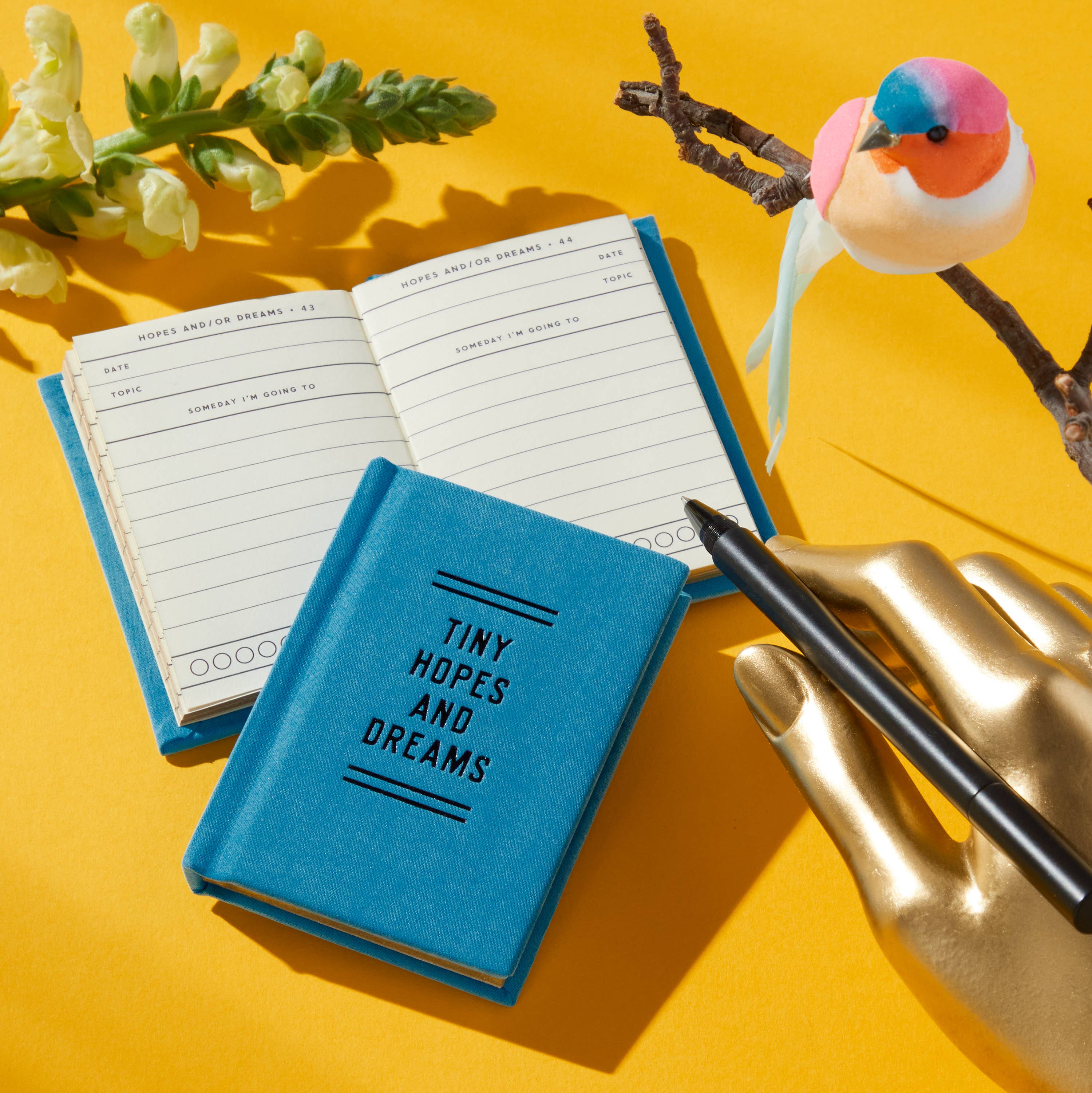 Blue 'Tiny Hopes and Dreams' journal with pen, golden hand, bird, and flowers on yellow background