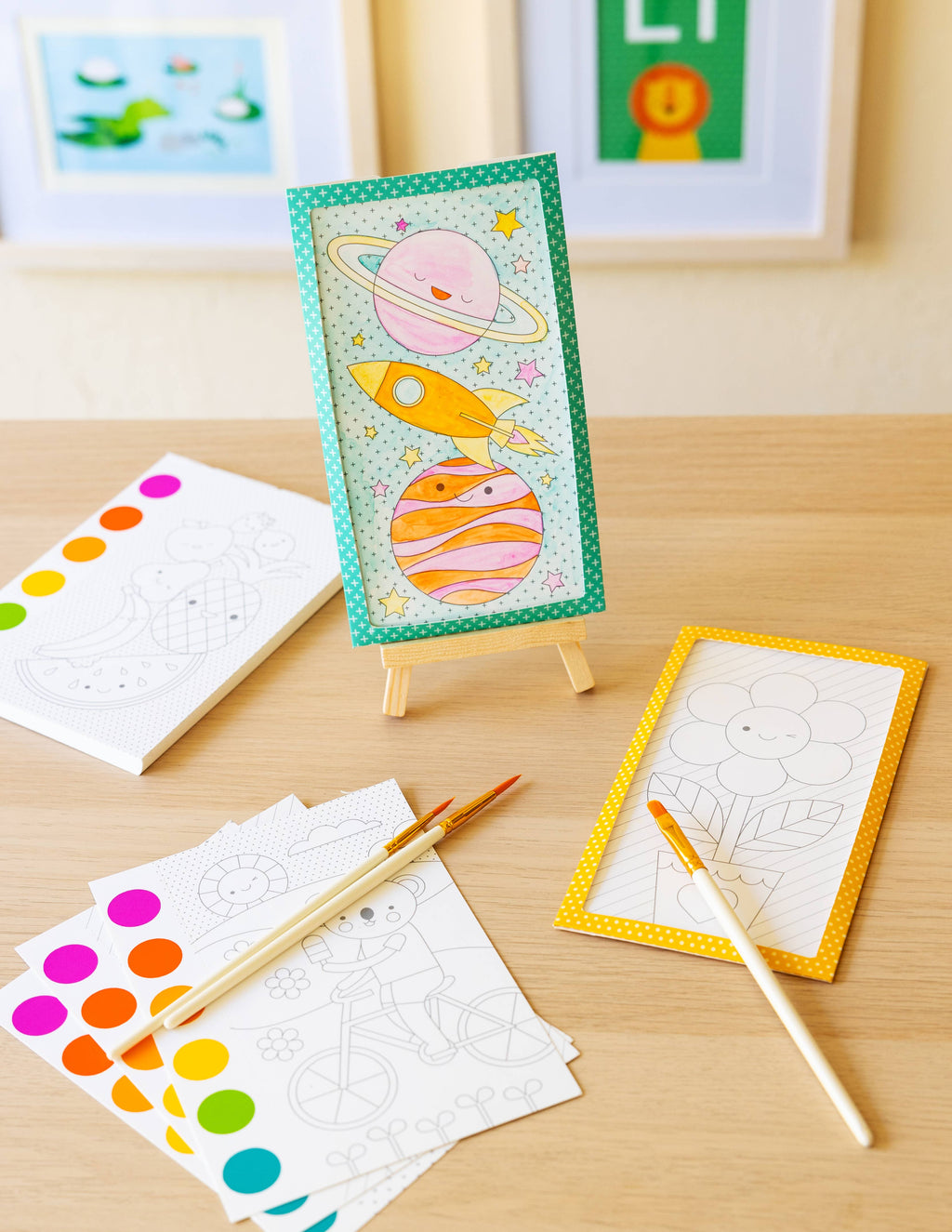 Children's art supplies including a drawing of planets and a rocket on an easel, with color swatches and brushes on a wooden surface.