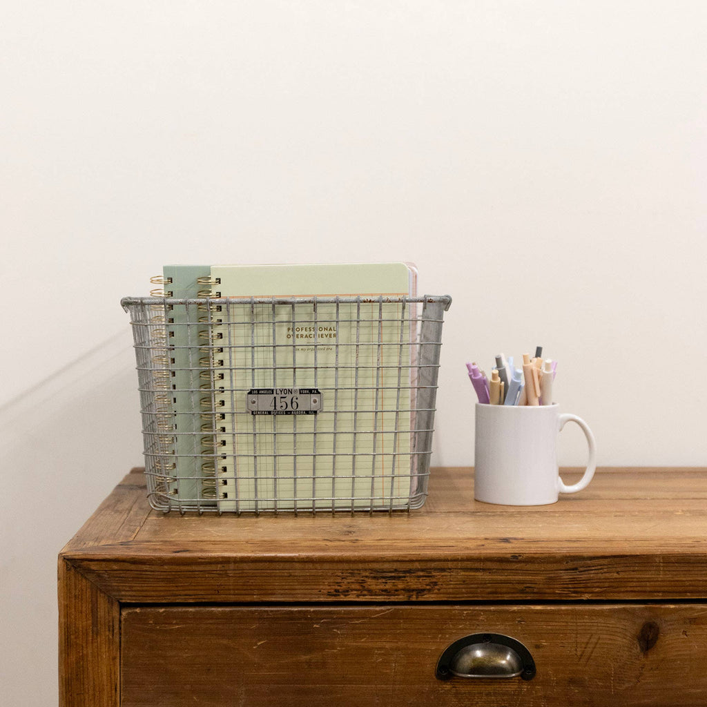 Metal wire basket with notebooks and a white mug with pens on a wooden surface.