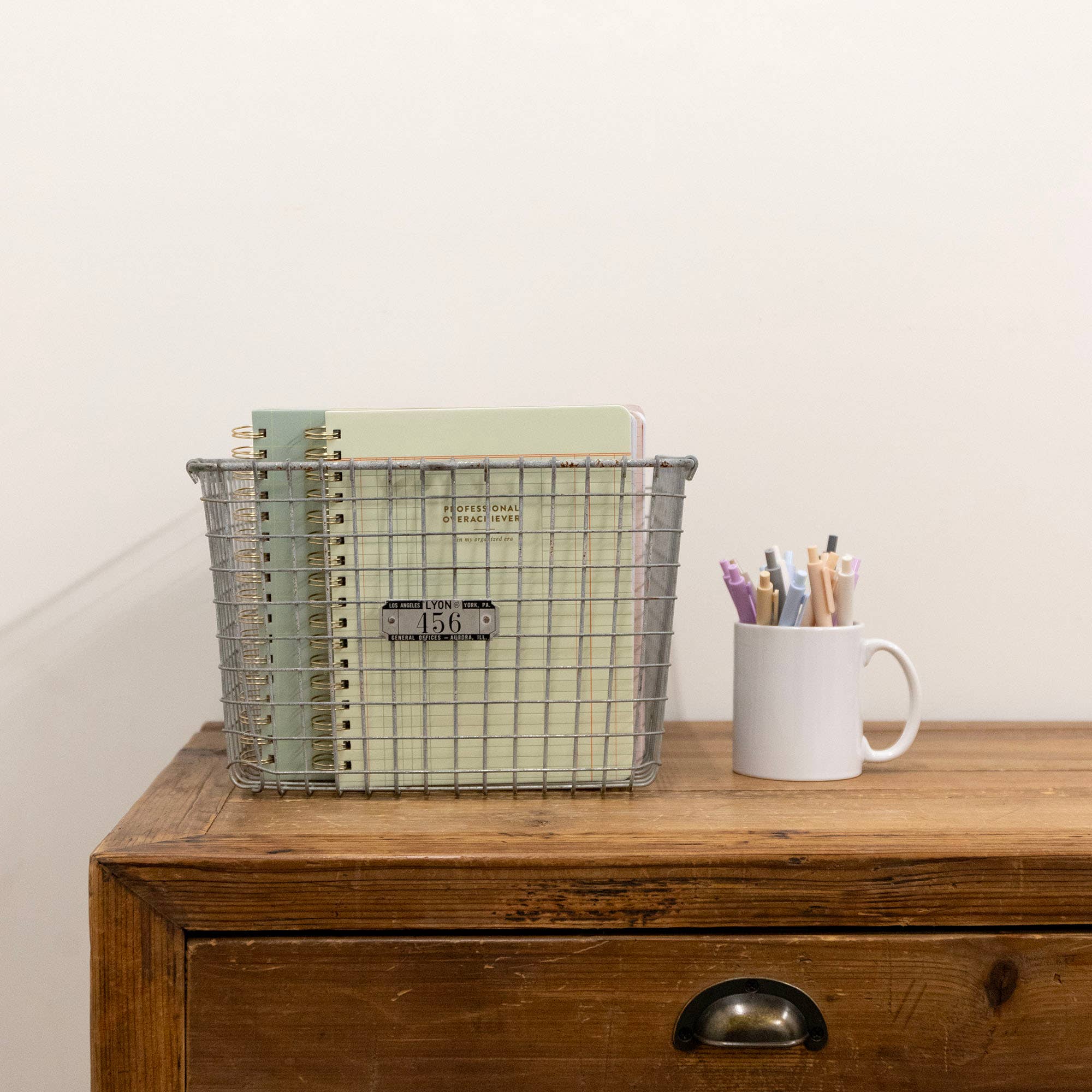 Metal wire basket with notebooks and a white mug with pens on a wooden surface.
