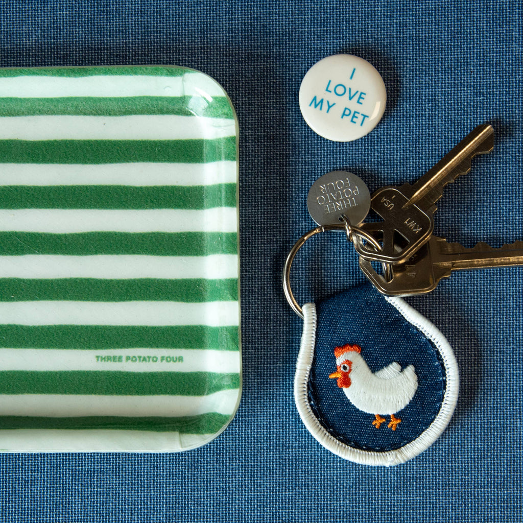 Keychain with chicken design, keys, and round tag on a striped tray against a blue background