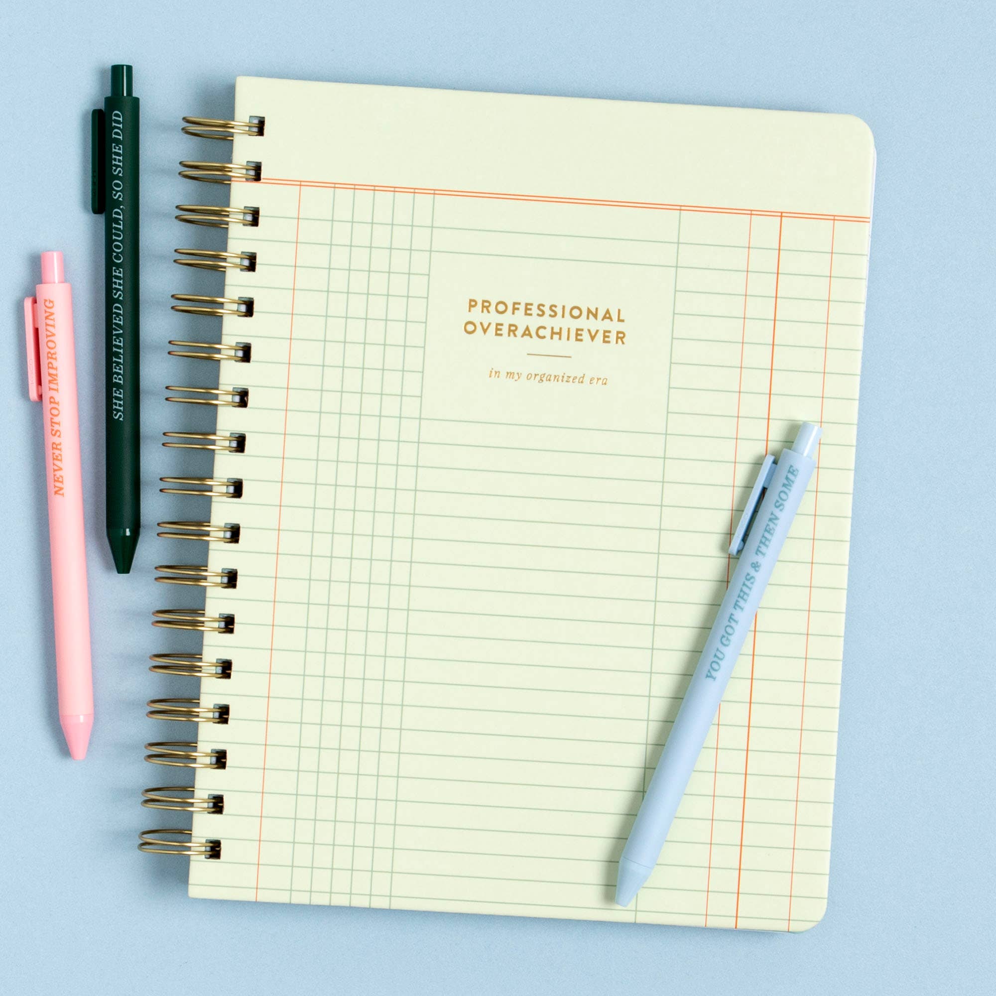 Notebook with grid pages and 'Professional Overachiever' text, surrounded by pens on a light blue background